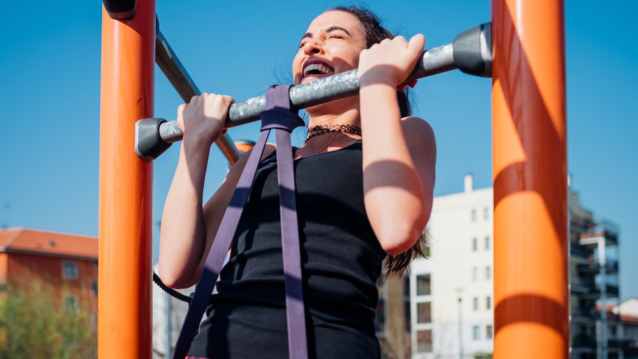 A photo of a woman wearing gym clothes completing a band assisted pull-up outdoors using a metal pull-up station.