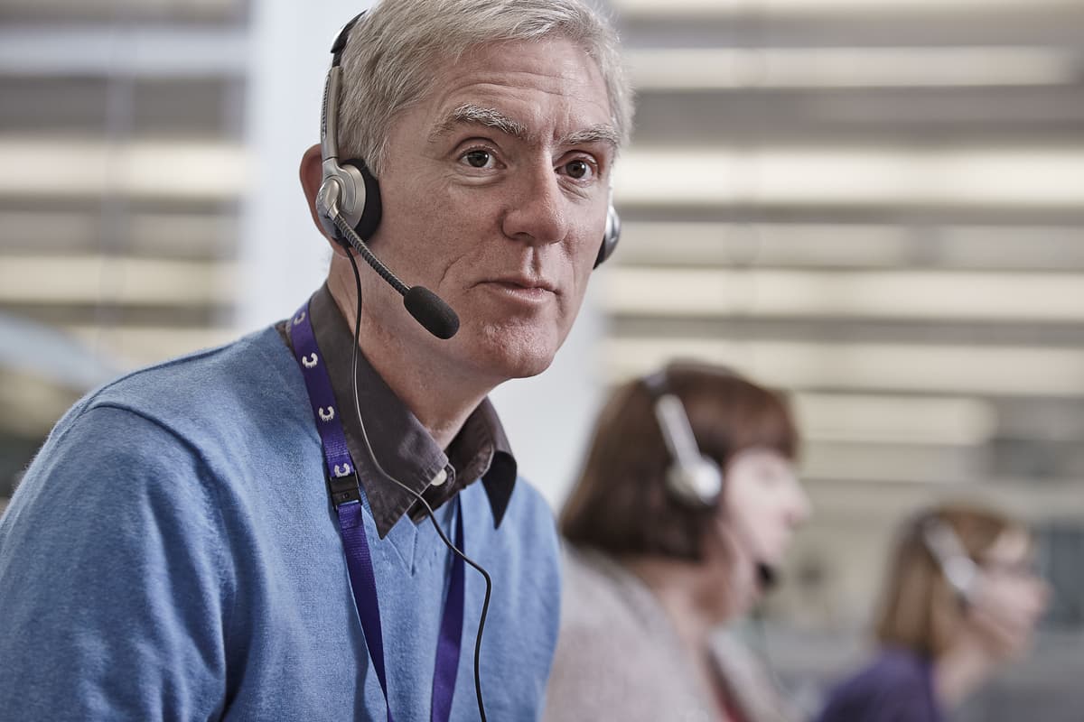 A man wearing a headset, speaking on a call. He appears to be working in an office as you can see others behind him.